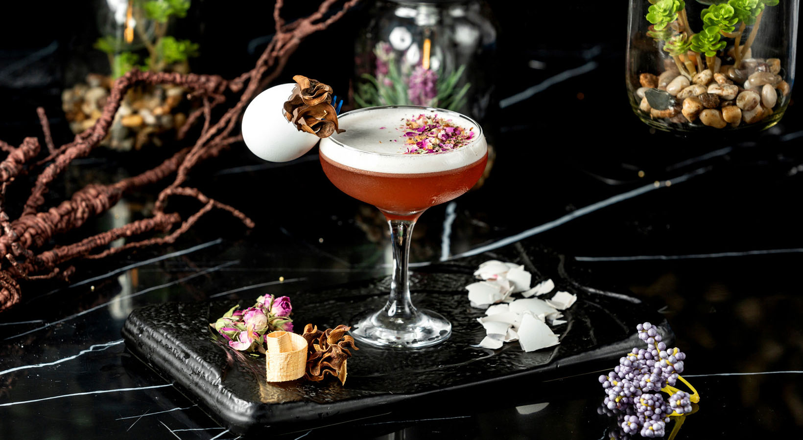 A cocktail sat on a black table with food and flower decorations around it